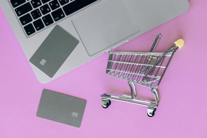 A miniature shopping cart on a pink surface next to a laptop.